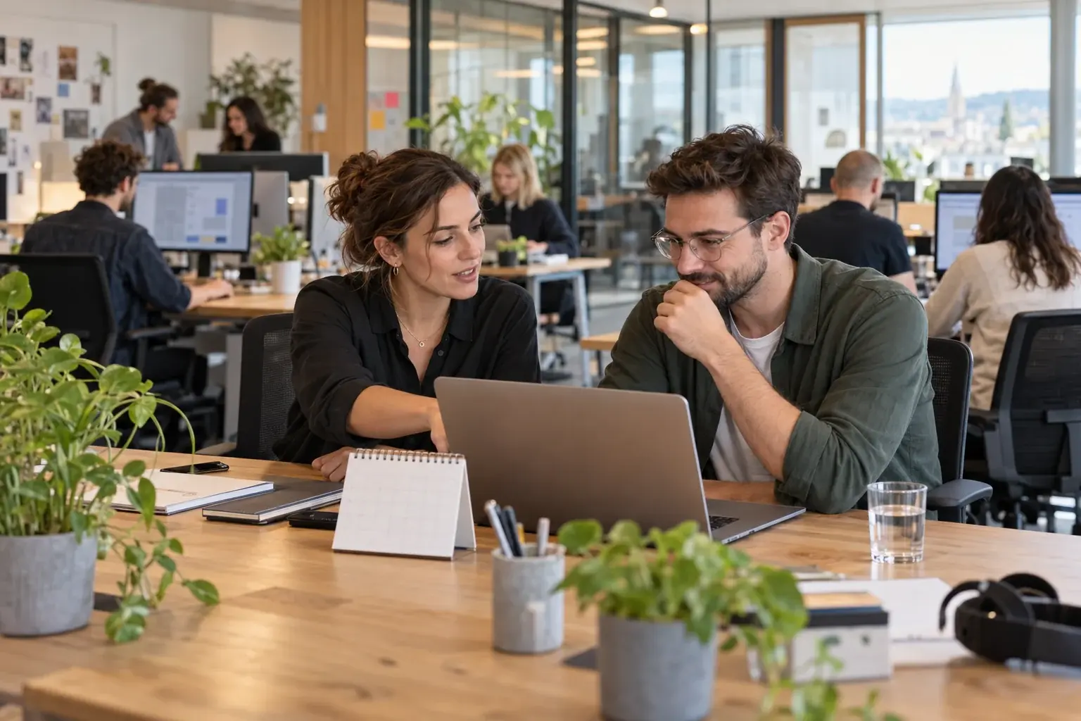 Zwei Personen besprechen die Teamplanung am Laptop; weitere Mitarbeitende arbeiten im offenen Büro.