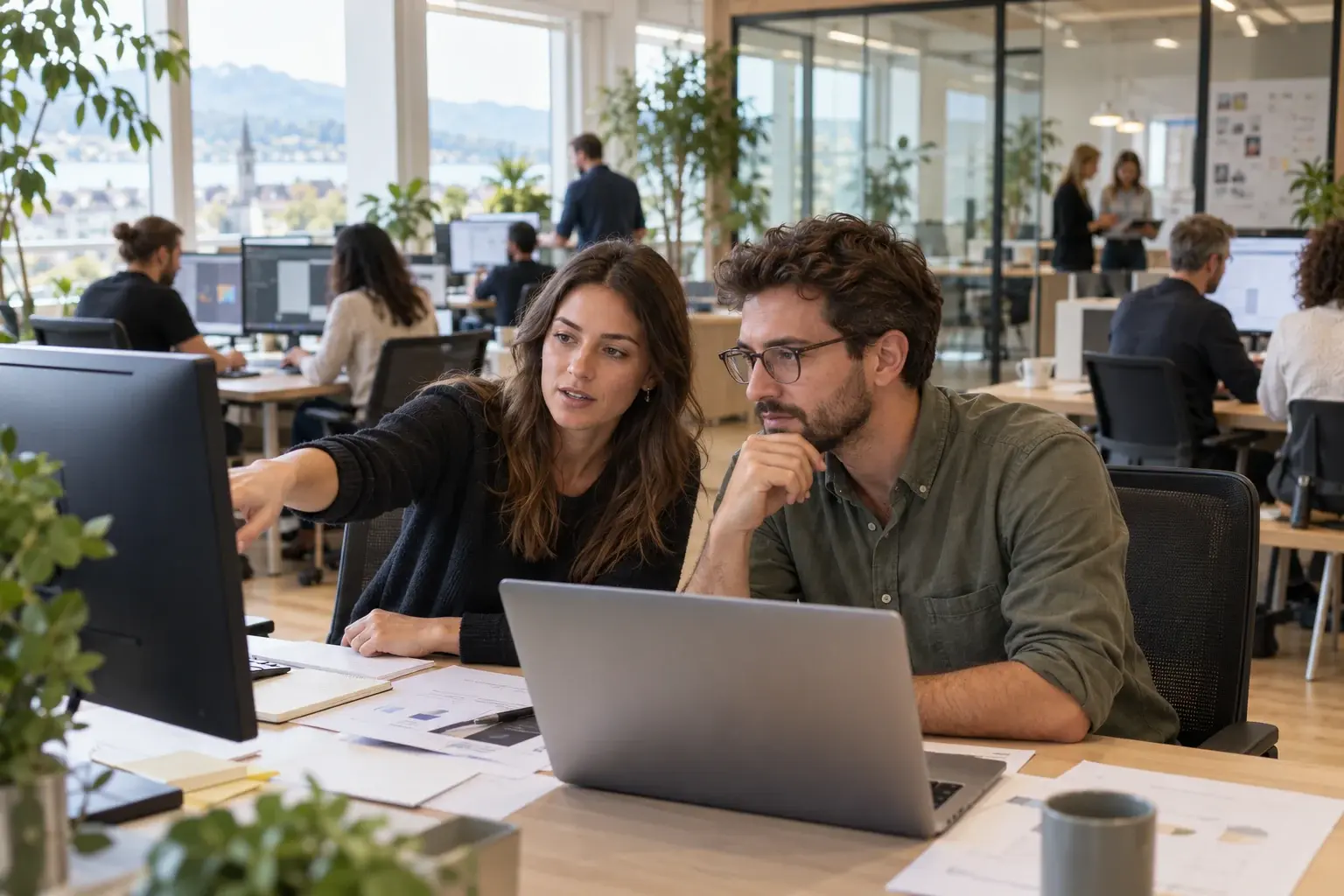 Zwei Personen besprechen einen Kundenauftrag am Bildschirm; im Hintergrund arbeitet ein Agenturteam.
